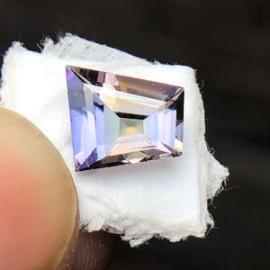May include: A rectangular, faceted gemstone with a purple and pink hue. The gemstone is set on a white surface.