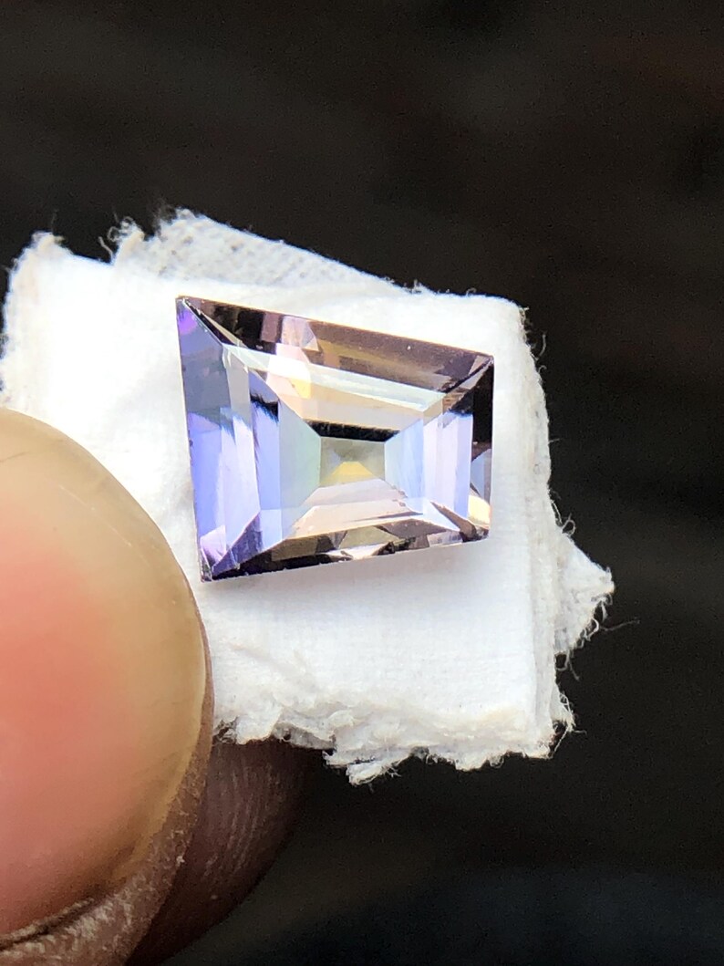 May include: A rectangular, faceted gemstone with a purple and pink hue. The gemstone is set on a white surface.