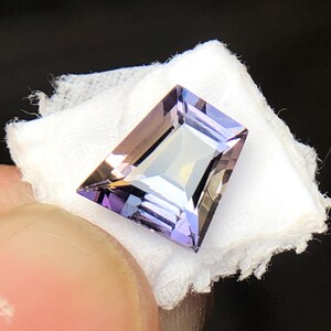 May include: A faceted, purple and brown gemstone with a rectangular shape. The gemstone is set on a white background.