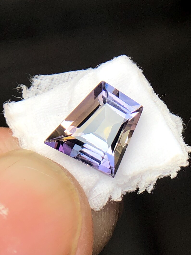 May include: A faceted, purple and brown gemstone with a rectangular shape. The gemstone is set on a white background.