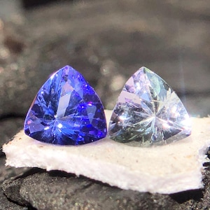 May include: Two triangular cut gemstones, one blue and one clear, are displayed on a white surface.