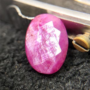 Peut inclure: Une pierre précieuse rubis de forme ovale à facettes, présentant une teinte magenta-rose vive. La pierre montre une structure interne visible en forme de grille et des inclusions naturelles sur toute sa surface, tenue par un outil métallique.