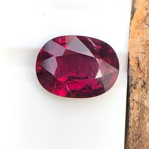 Puede incluir: Una piedra preciosa ovalada de color rojo intenso con facetas. La piedra preciosa está sobre una superficie blanca.
