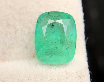 May include: A vivid green cushion-cut emerald gemstone. The gem has visible inclusions and facets that reflect light. The emerald is set against a white, textured background, enhancing its colour and clarity. A beautiful gemstone for jewellery.