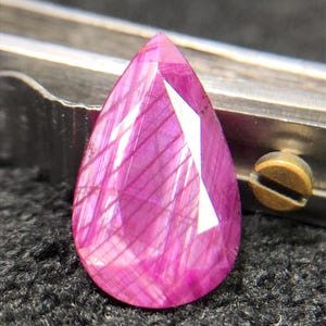 Op de afbeelding: Een peervormige, gefacetteerde edelsteen met een levendige magenta-roze tint. De steen vertoont duidelijke interne lineaire insluitingen en een gepolijst oppervlak dat licht reflecteert over de vele hoekige facetten van de steen.