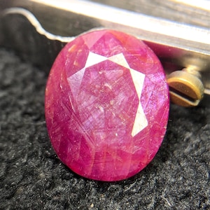 Puede incluir: Una piedra preciosa de corte ovalado en color rosa magenta vibrante con una superficie facetada. La piedra muestra inclusiones cristalinas internas y facetas que reflejan la luz, sujeta por una pinza metálica con un tornillo visible.