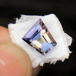 May include: A faceted, rectangular, purple gemstone with a light blue hue. The gemstone is set on a white fabric background.