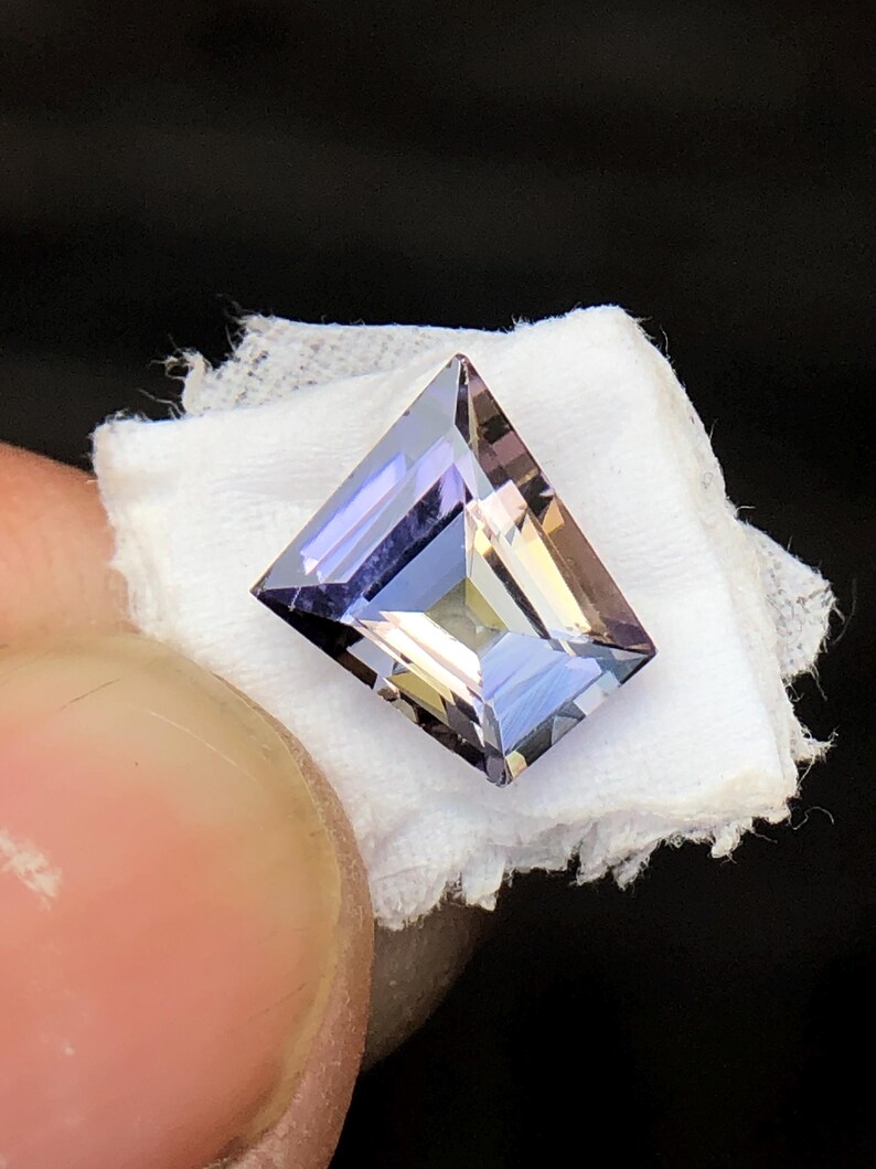 May include: A faceted, rectangular, purple gemstone with a light blue hue. The gemstone is set on a white fabric background.