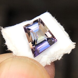 May include: A faceted, rectangular amethyst gemstone with a purple and brown hue. The gemstone is set on a white background.