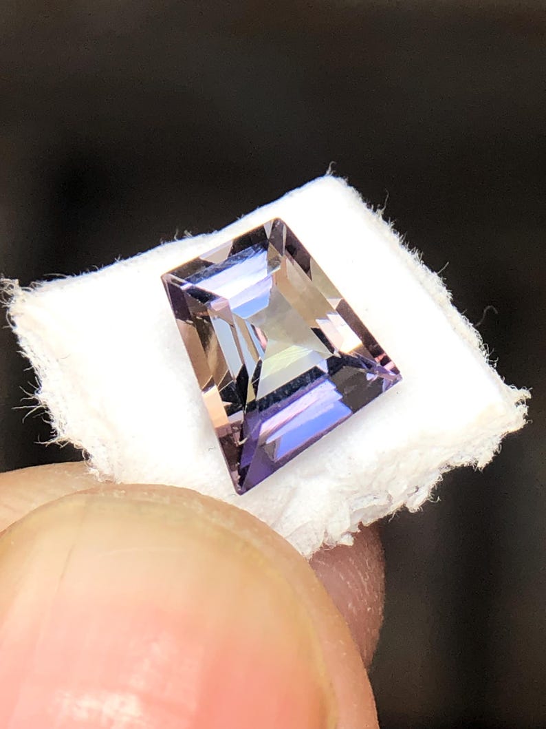 May include: A faceted, rectangular amethyst gemstone with a purple and brown hue. The gemstone is set on a white background.