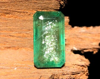 May include: A rectangular-cut emerald gemstone with a vibrant green hue. The gem is set against a textured, brown wooden surface, with light reflecting off its facets. The emerald's clarity is visible, showcasing its natural beauty.