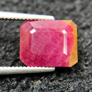May include: A rectangular faceted gemstone with a deep red centre that transitions into a translucent orange-yellow hue along the right edge. The stone is held by silver-toned metal tweezers, showcasing its polished surface and internal inclusions.