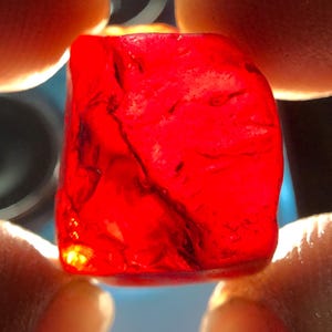 Könnte beinhalten: Ein lebendiger, durchscheinender roter Edelstein, der zwischen zwei Fingern gehalten wird. Der Stein hat eine raue, unregelmäßige Form mit inneren Adern und leuchtet mit einem inneren Licht. Die Farbe des Edelsteins ist ein tiefes, sattes Rot.