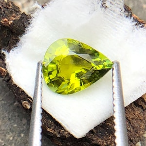 Op de afbeelding: Een peervormige groene edelsteen met een facetgeslepen vorm, vastgehouden door zilveren pincetten op een wit wattenpad.