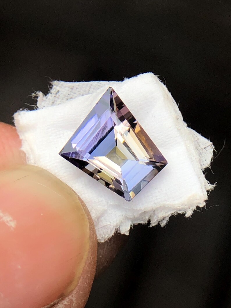 May include: A faceted, rectangular, purple and brown gemstone with a light, almost translucent appearance.