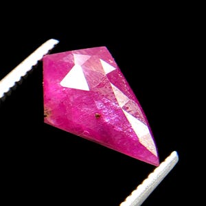 May include: A kite-shaped, faceted gemstone with a vibrant magenta-pink hue. The stone features a geometric cut with multiple flat surfaces that reflect light, creating bright white highlights across its translucent, crystalline structure.