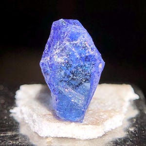 May include: A striking blue crystal with a textured surface. It is mounted on a small, off-white, rectangular base. The dark background accentuates the crystal's colour and shape. The crystal appears to be a natural mineral.