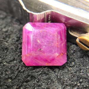 Puede incluir: Una piedra preciosa de color magenta vibrante con corte cuadrado y superficies facetadas, que muestra inclusiones internas y variaciones naturales en la intensidad del color, colocada bajo la punta metálica de unas pinzas de precisión.