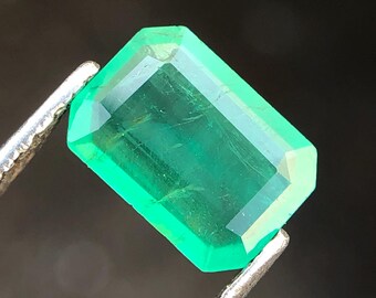 May include: A vibrant, emerald-cut emerald gemstone, held by silver tweezers. The gem is a vivid green color with a rectangular shape and faceted edges, showcasing its clarity and brilliance. The background is a dark, blurred backdrop.
