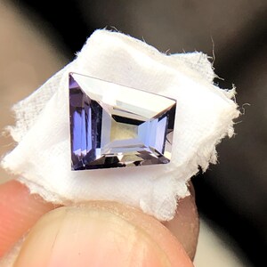 May include: A purple gemstone with a rectangular shape, cut into a faceted design. The gemstone is set on a white background.