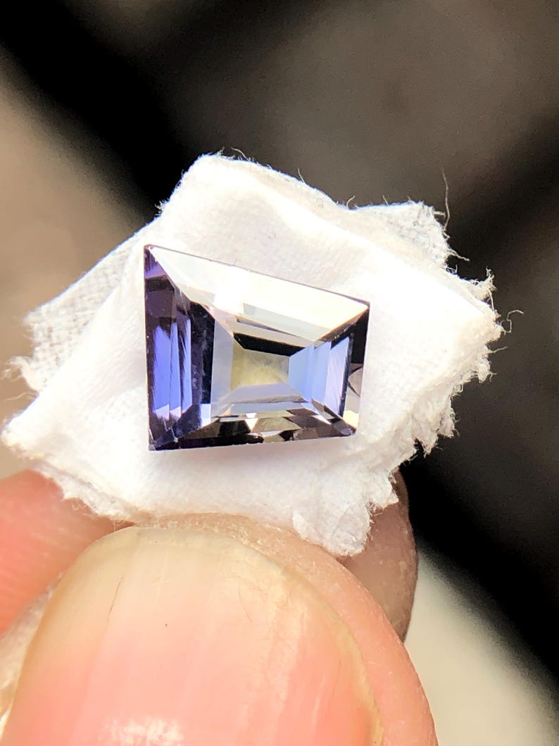 May include: A purple gemstone with a rectangular shape, cut into a faceted design. The gemstone is set on a white background.