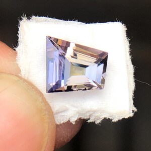 May include: A light purple gemstone with a square shape and a faceted cut. The gemstone is displayed on a white fabric background.