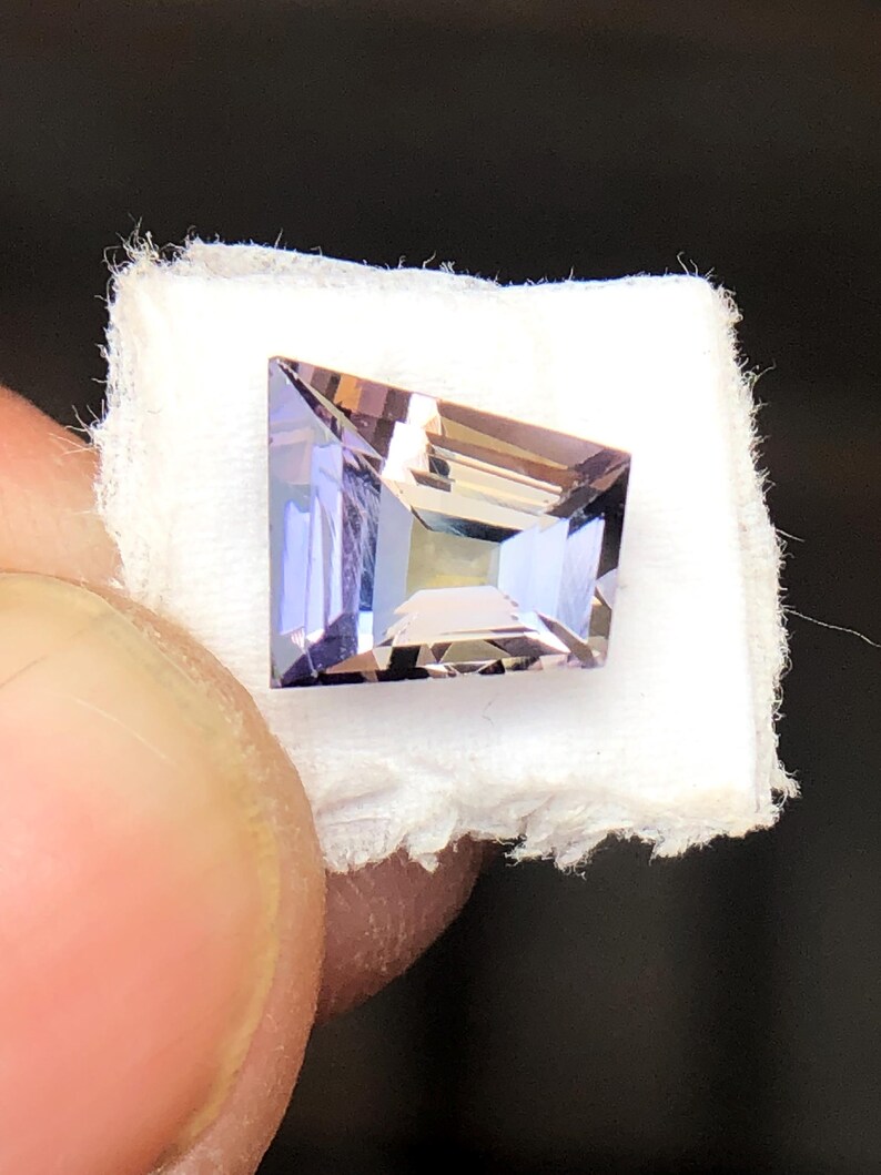 May include: A light purple gemstone with a square shape and a faceted cut. The gemstone is displayed on a white fabric background.