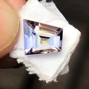 May include: A light purple gemstone with a rectangular shape and a faceted cut. The gemstone is set on a white background.