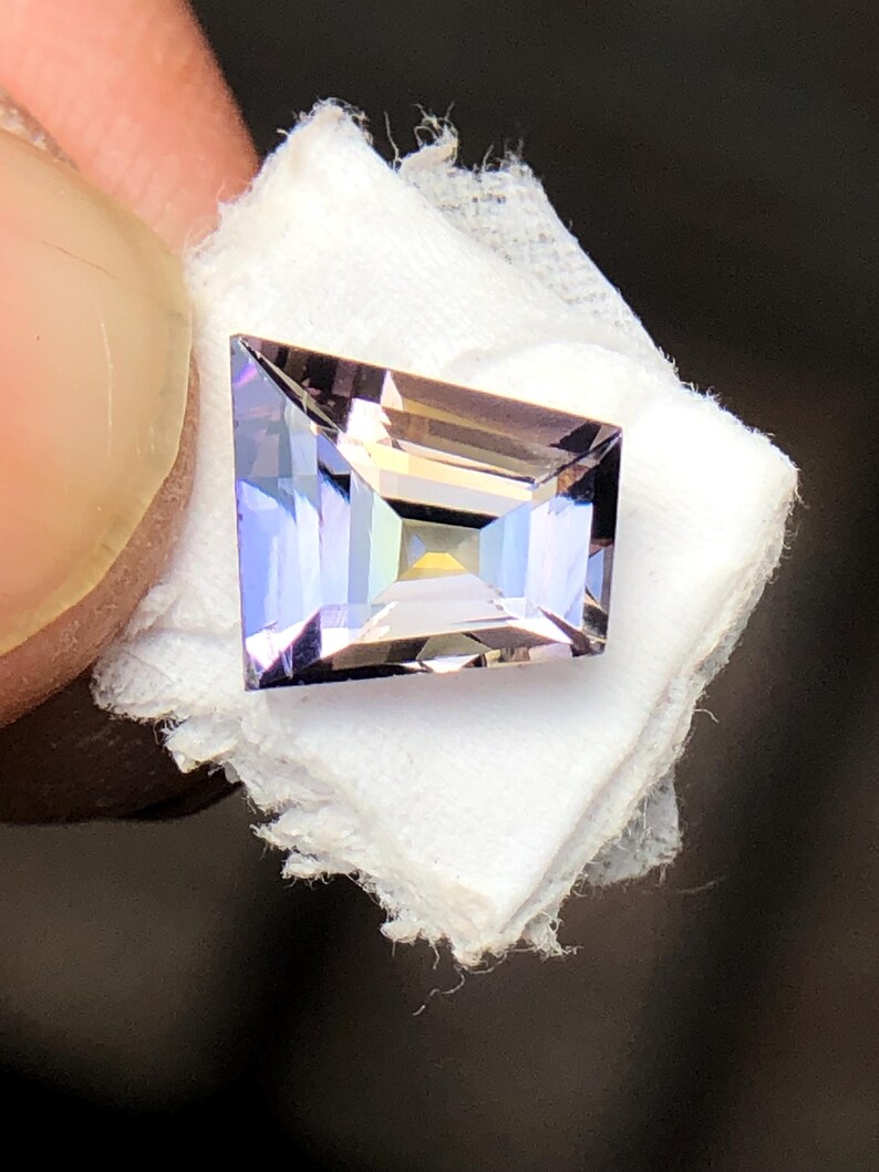 May include: A light purple gemstone with a rectangular shape and a faceted cut. The gemstone is set on a white background.