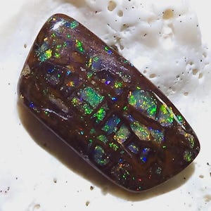 Op de afbeelding: Een gepolijste, rechthoekige edelsteen met een donkerbruine basis en een mozaïek van iriserende kleuren. De edelsteen heeft flitsen van groen, blauw en goud, wat een levendige weergave creëert. De steen ligt tegen een witte achtergrond.