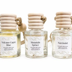 May include: Three clear glass car air freshener bottles with light-colored wooden caps and hanging jute cords. Each bottle has a white label with black text reading Volcano Capri Blue, Mountain Spruce, or Narcissist, plus Net Wt. 22 oz (59 g).