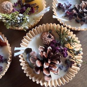 Puede incluir: Primer plano de varias velas pequeñas y redondas en moldes de papel. Cada vela está decorada con elementos naturales como piñas, flores secas y ramitas. Las velas tienen un aspecto rústico y hecho a mano, ideal para la decoración del hogar o regalos.
