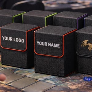 May include: Black card storage boxes with colored trim and custom text options. Some boxes have the text "YOUR LOGO" and "YOUR NAME" in white. One box features a gold emblem. The boxes are on a patterned surface.