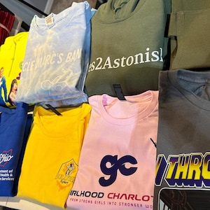May include: A collection of folded t-shirts and hoodies in various colors, including blue, yellow, pink, and green. Some shirts feature text like "CLE MARC'S BAN", "Ohio Department of Mental Health & Addiction Services", and "Girlhood Charlotte".