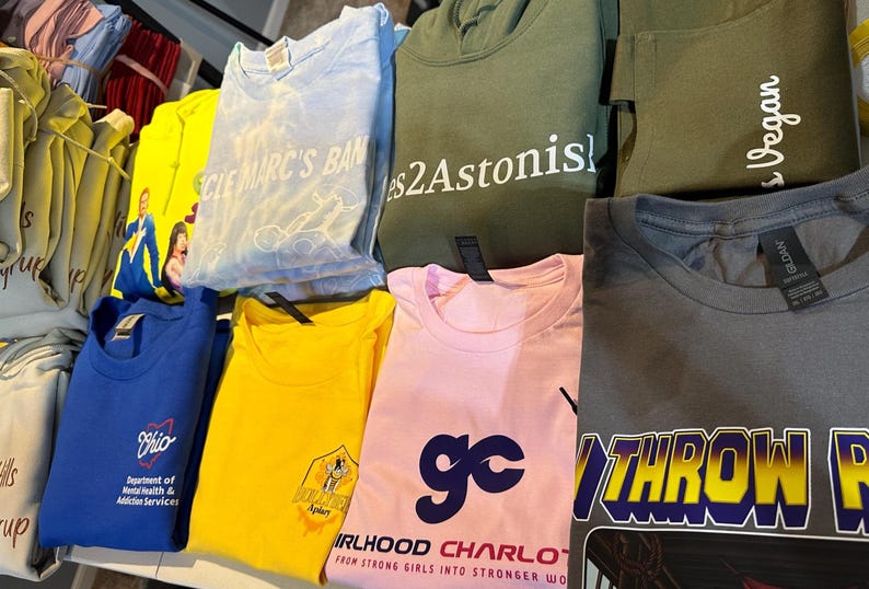 May include: A collection of folded t-shirts and hoodies in various colors, including blue, yellow, pink, and green. Some shirts feature text like "CLE MARC'S BAN", "Ohio Department of Mental Health & Addiction Services", and "Girlhood Charlotte".