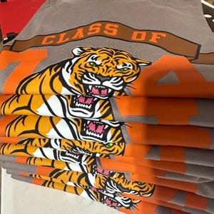 May include: A stack of gray t-shirts featuring a tiger graphic with orange and white stripes, green eyes, and a pink mouth. The shirts have the text "CLASS OF 74" in orange and brown.