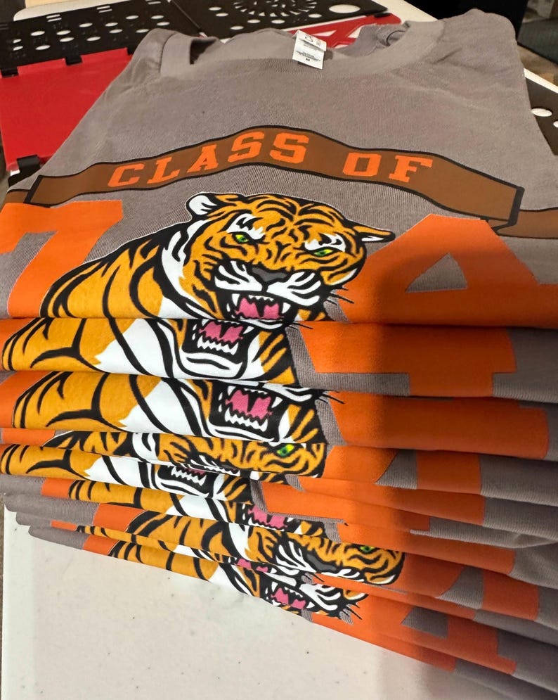 May include: A stack of gray t-shirts featuring a tiger graphic with orange and white stripes, green eyes, and a pink mouth. The shirts have the text "CLASS OF 74" in orange and brown.