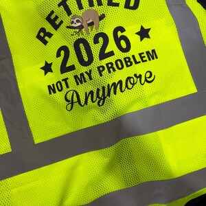 Op de afbeelding: Een neongeel veiligheidsvest met hoge zichtbaarheid en zwarte tekst RETIRED 2026 NOT MY PROBLEM Anymore, inclusief een afbeelding van een bruine luiaard op een tak. Het vest heeft horizontale en verticale grijze reflecterende strepen.