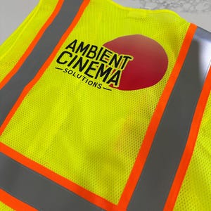 Op de afbeelding: Een neongeel veiligheidsvest met reflecterende zilveren strepen en feloranje randen. Op de achterkant staat een rode cirkelvormige afbeelding met de zwarte tekst AMBIENT CINEMA SOLUTIONS in een vetgedrukt, schreefloos lettertype.