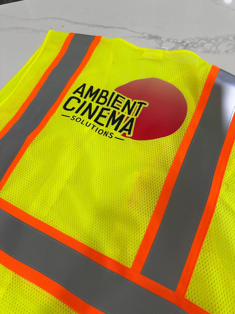 Op de afbeelding: Een neongeel veiligheidsvest met reflecterende zilveren strepen en feloranje randen. Op de achterkant staat een rode cirkelvormige afbeelding met de zwarte tekst AMBIENT CINEMA SOLUTIONS in een vetgedrukt, schreefloos lettertype.