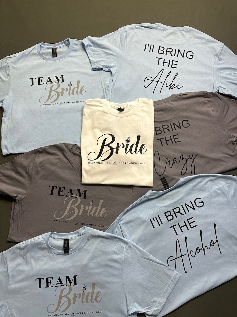 May include: A collection of light blue, gray, and white t-shirts. Some shirts say "Team Bride" with the location and date. Other shirts say "I'll Bring the Alibi", "Bring the Crazy", and "I'll Bring the Alcohol".