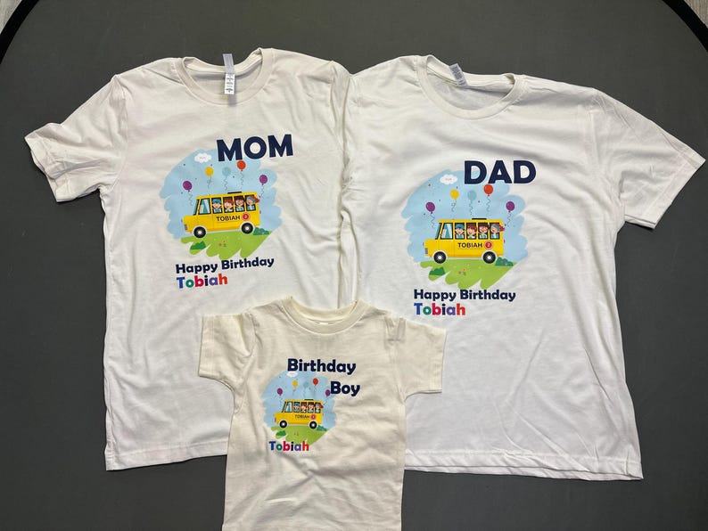 May include: White t-shirts with custom designs for a family. The shirts feature a yellow school bus graphic with the text "Happy Birthday Tobiah". One shirt says "MOM", another says "DAD", and the third says "Birthday Boy".