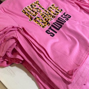 May include: A stack of pink t-shirts. Each shirt features the text "JUST DANCE STUDIO 55" in a combination of leopard print and black lettering. The shirts are neatly folded, suggesting they are ready for sale or distribution.
