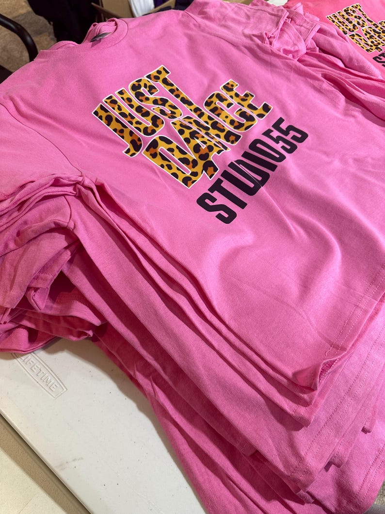 May include: A stack of pink t-shirts. Each shirt features the text "JUST DANCE STUDIO 55" in a combination of leopard print and black lettering. The shirts are neatly folded, suggesting they are ready for sale or distribution.