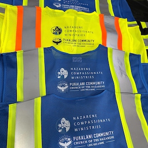 Op de afbeelding: Een collectie veiligheidsvesten met hoge zichtbaarheid in blauw en neongeel, voorzien van reflecterende zilveren strepen. Elk vest is bedrukt met de tekst Nazarene Compassionate Ministries en Pukalani Community Church of the Nazarene Love Has a Name.
