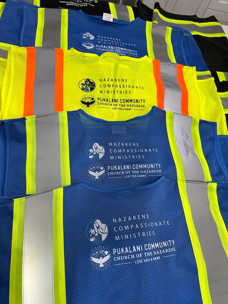 Op de afbeelding: Een collectie veiligheidsvesten met hoge zichtbaarheid in blauw en neongeel, voorzien van reflecterende zilveren strepen. Elk vest is bedrukt met de tekst Nazarene Compassionate Ministries en Pukalani Community Church of the Nazarene Love Has a Name.