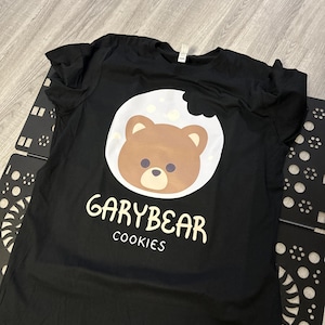 May include: Black t-shirt featuring a brown bear face inside a white cookie with a bite taken out. The text "GARYBEAR COOKIES" is printed below the bear graphic in white. The shirt is laying on a light gray surface.