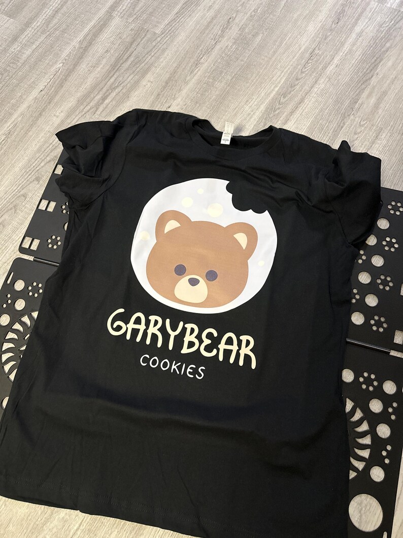 May include: Black t-shirt featuring a brown bear face inside a white cookie with a bite taken out. The text "GARYBEAR COOKIES" is printed below the bear graphic in white. The shirt is laying on a light gray surface.