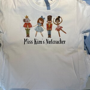 May include: White t-shirt featuring a colorful graphic of characters from The Nutcracker ballet, including a mouse king, a ballerina, and a nutcracker soldier. The text "Miss Kim's Nutcracker" is printed below the characters.