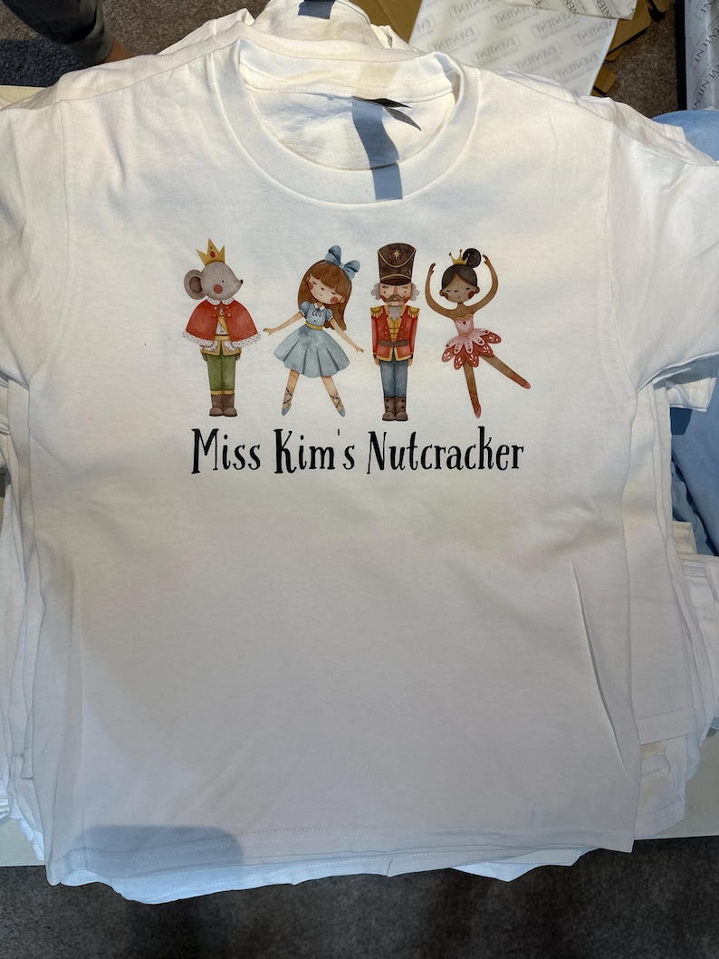 May include: White t-shirt featuring a colorful graphic of characters from The Nutcracker ballet, including a mouse king, a ballerina, and a nutcracker soldier. The text "Miss Kim's Nutcracker" is printed below the characters.